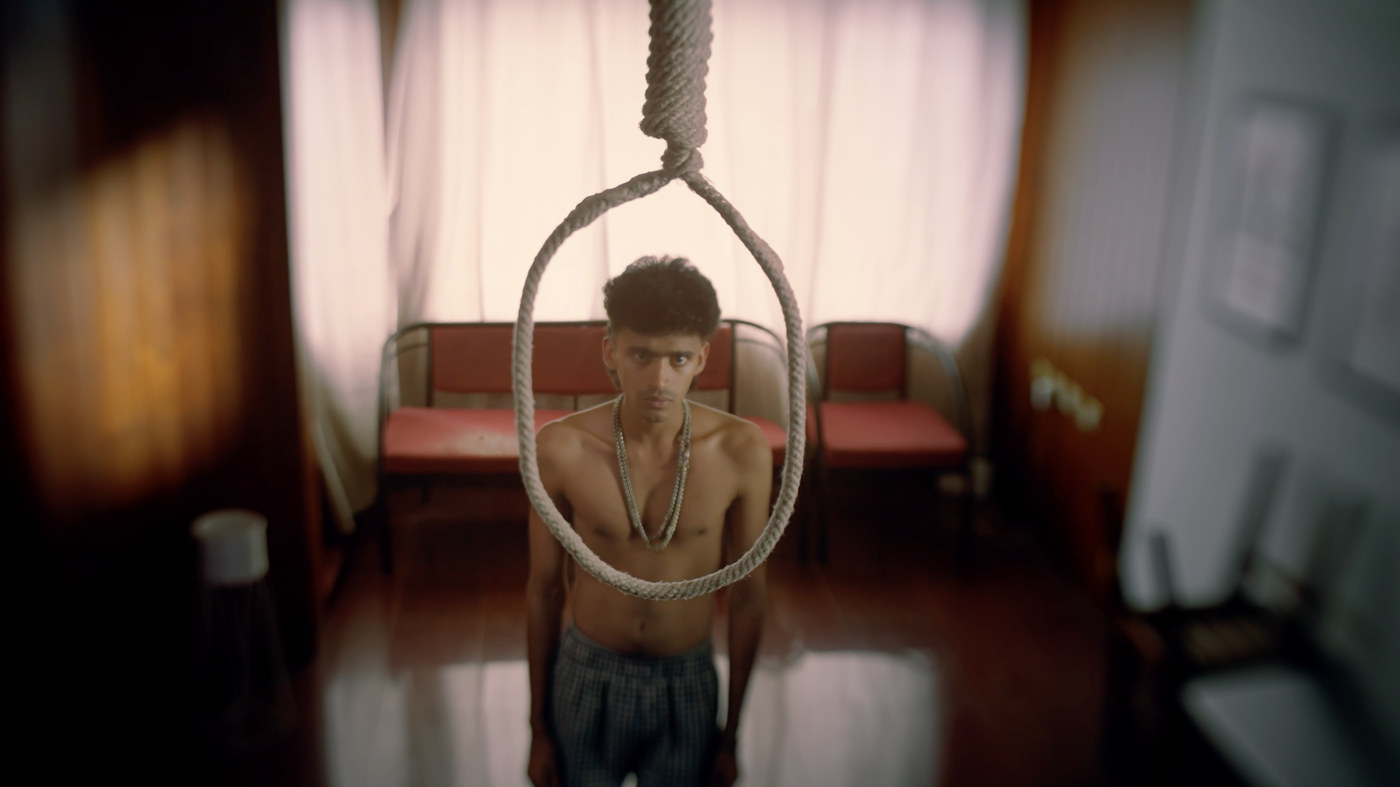 A shirtless man standing beneath a hanging noose in a dim room
